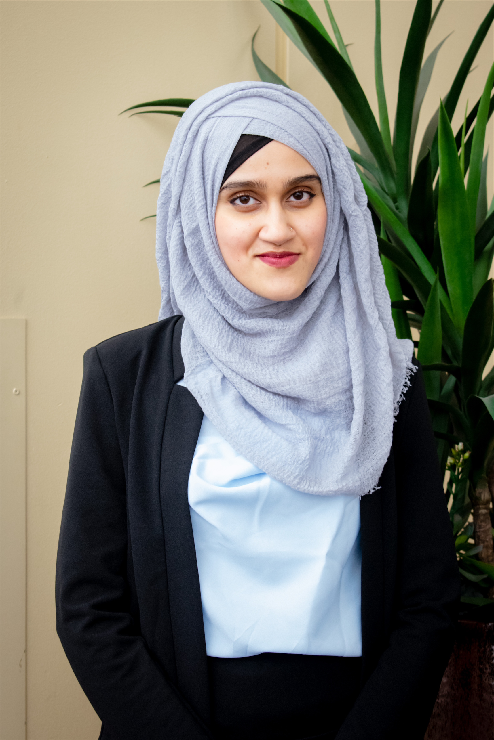 A professional photo of Nabila Ali standing, wearing a hijab and a suit, standing in front of a plant.